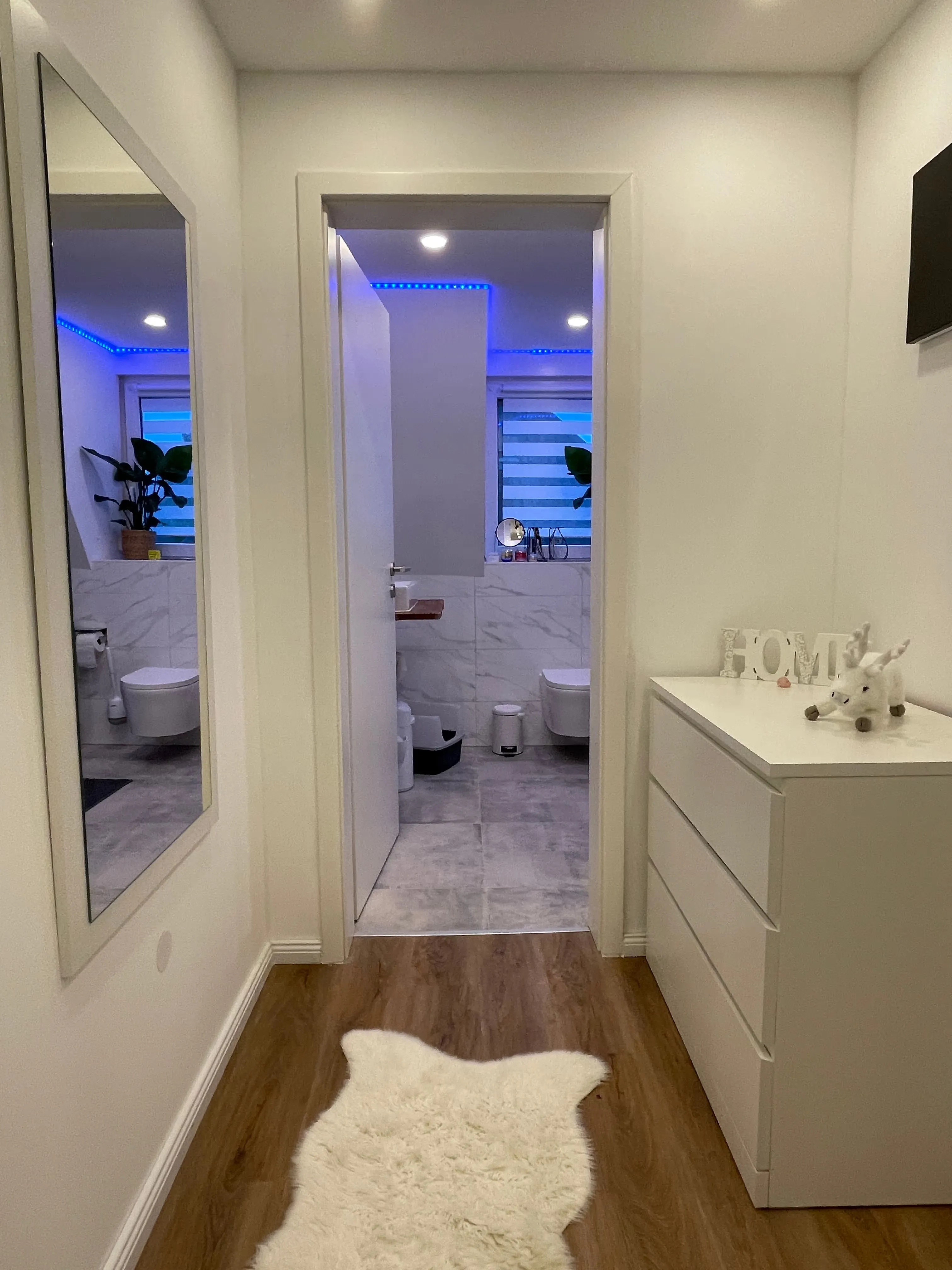 Flur mit holzfarbenem Boden, weißem Fellteppich, großer rechteckiger Wandspiegel links und weißer Kommode rechts mit Dekoration, Blick in ein Badezimmer mit weißen Fliesen und blauer LED-Beleuchtung.