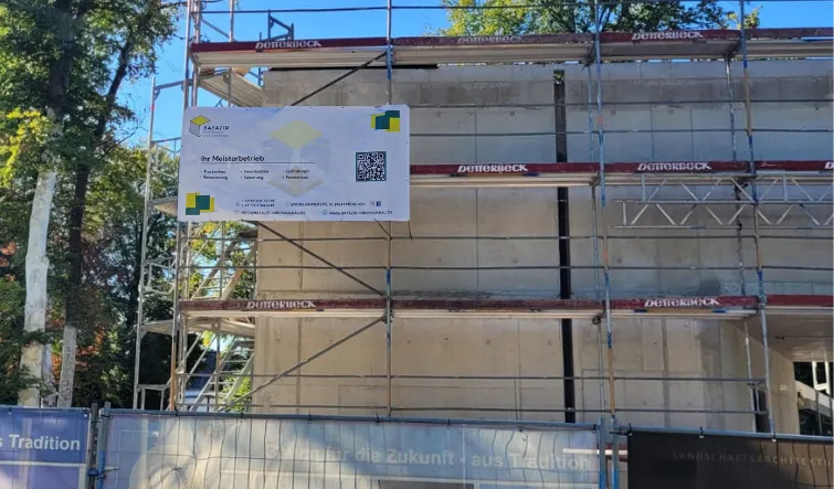 Baugerüst vor einem unvollendeten Gebäude mit einem Baustellenschild und Bäumen im Hintergrund.
