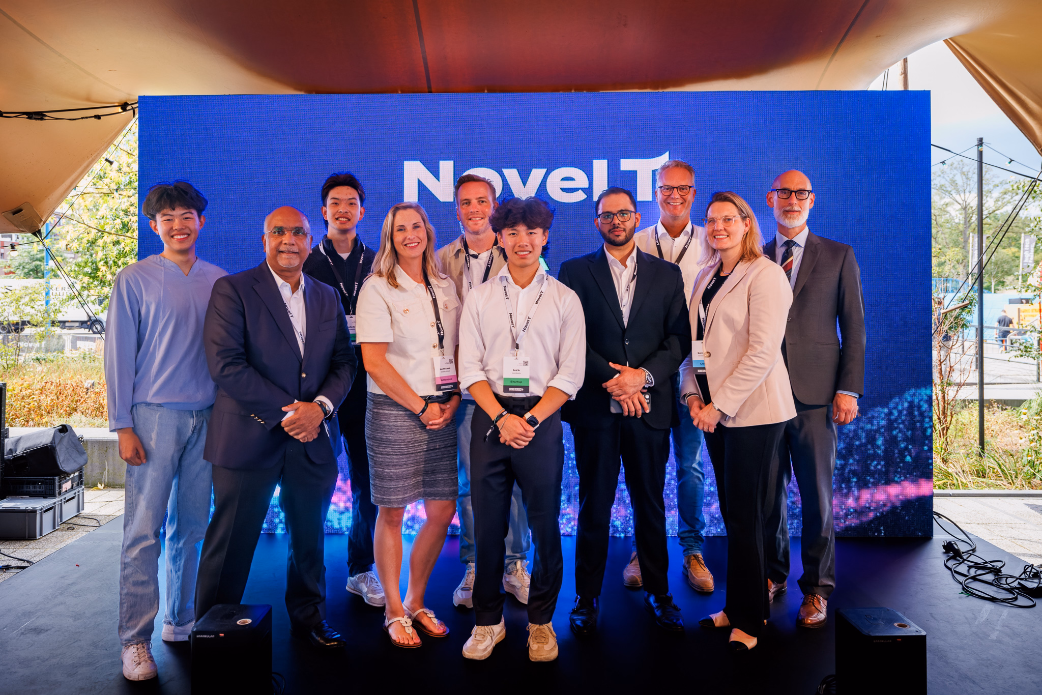 Group of nine people standing together and smiling at an event, posing in front of a large blue screen with the text ‘Novel-T’ and 'Velocity' displayed.