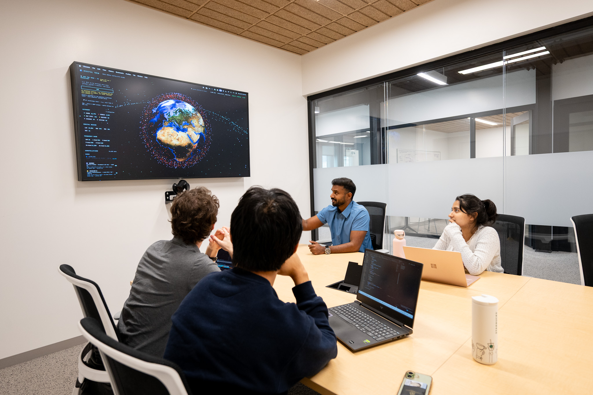 Photon-IV team members sitting around a meeting room table, collaborating on a SATCOM challenge while viewing a large screen displaying satellites positioned around the globe.