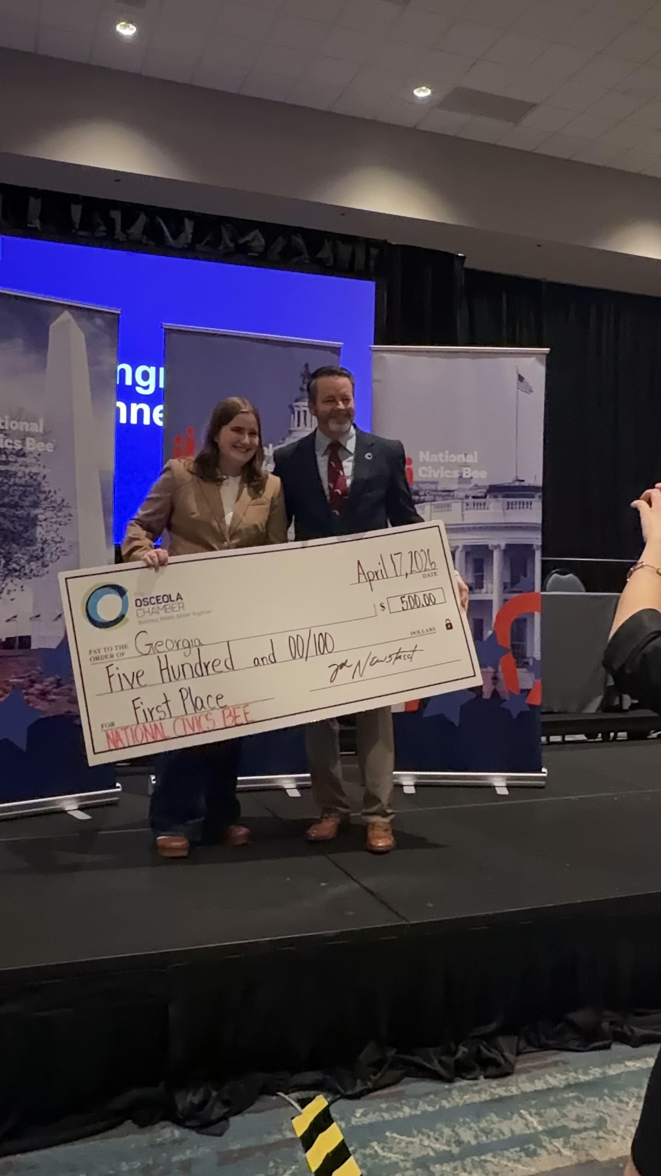 First-place winner holds an oversized prize check on stage at the 2026 Osceola Chamber Regional Civics Bee.
