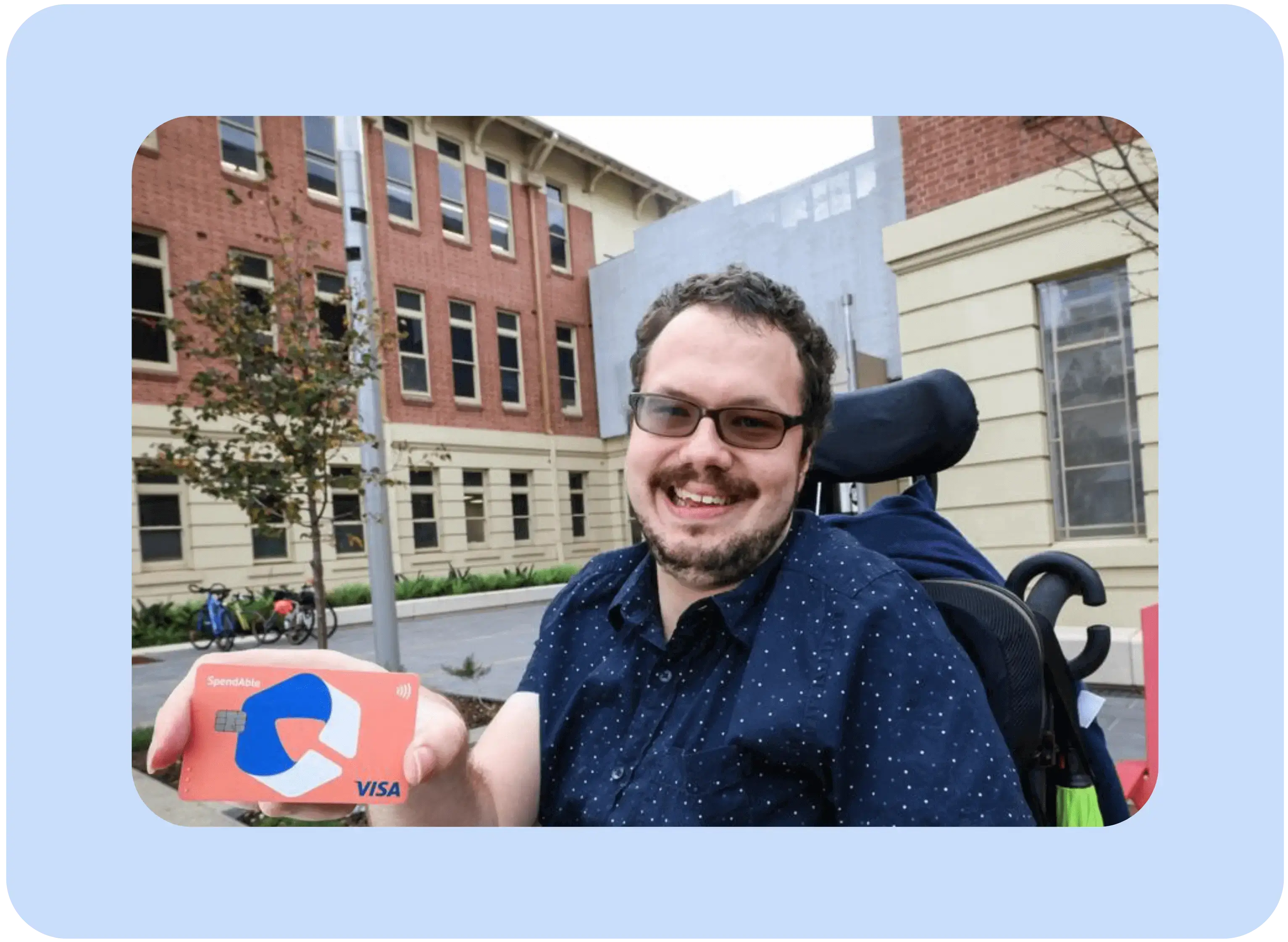 A smiling man in a wheelchair holds a SpendAble Visa card outdoors, representing financial independence and accessibility through the SpendAble money management app.