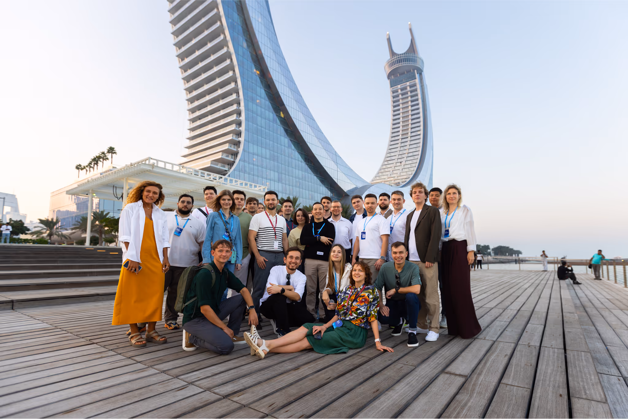 Group of diverse people posing on a wooden boardwalk with a modern curved skyscraper and palm trees in the background.