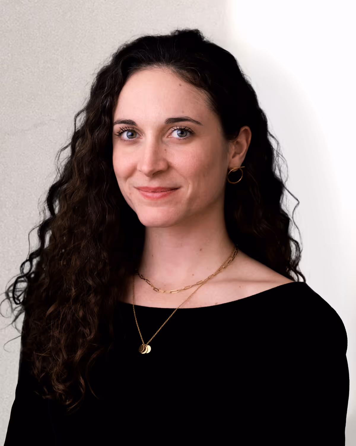 Woman with long curly dark hair wearing a black top, gold hoop earrings, and layered gold necklaces, smiling softly.