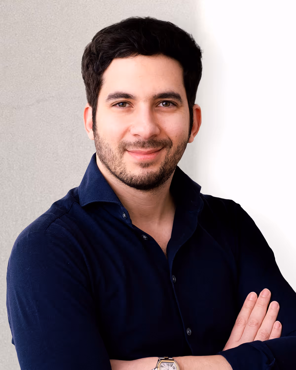 Smiling man with short dark hair and beard wearing a dark blue shirt and a watch, standing with arms crossed.