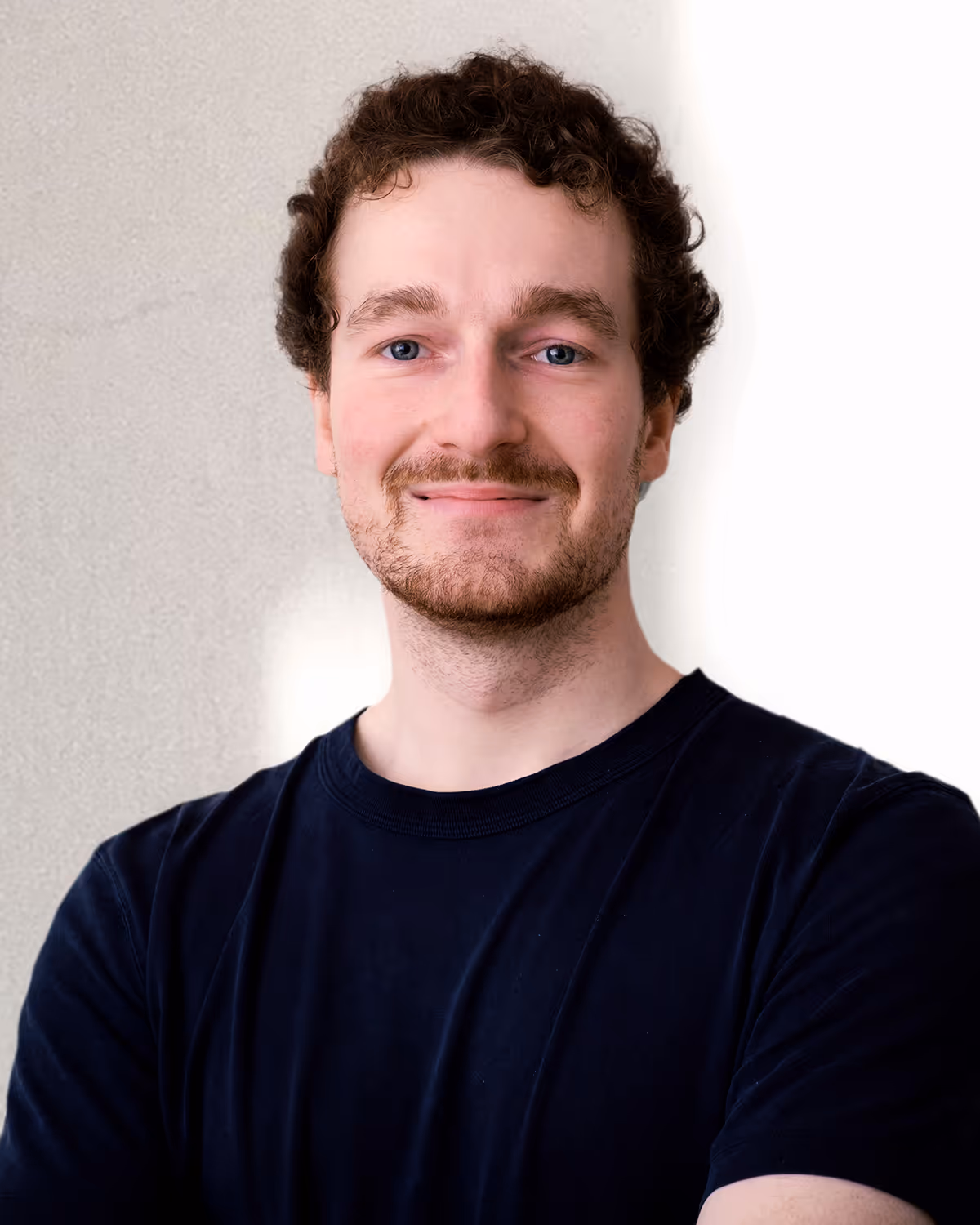 Smiling young man with curly brown hair and beard wearing a black t-shirt, standing against a neutral background.