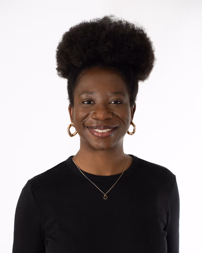 Smiling woman with natural curly hair wearing gold hoop earrings, a delicate necklace, and a black top against a white background.