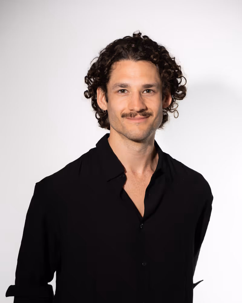 Man with curly hair, a mustache, and an earring wearing a black button-up shirt against a white background.
