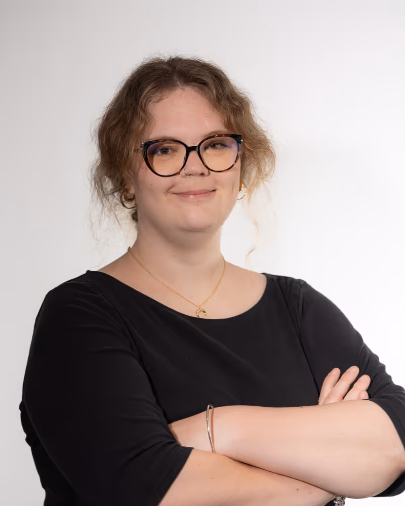 Smiling woman with glasses, curly hair, and crossed arms wearing a black top and gold jewelry.