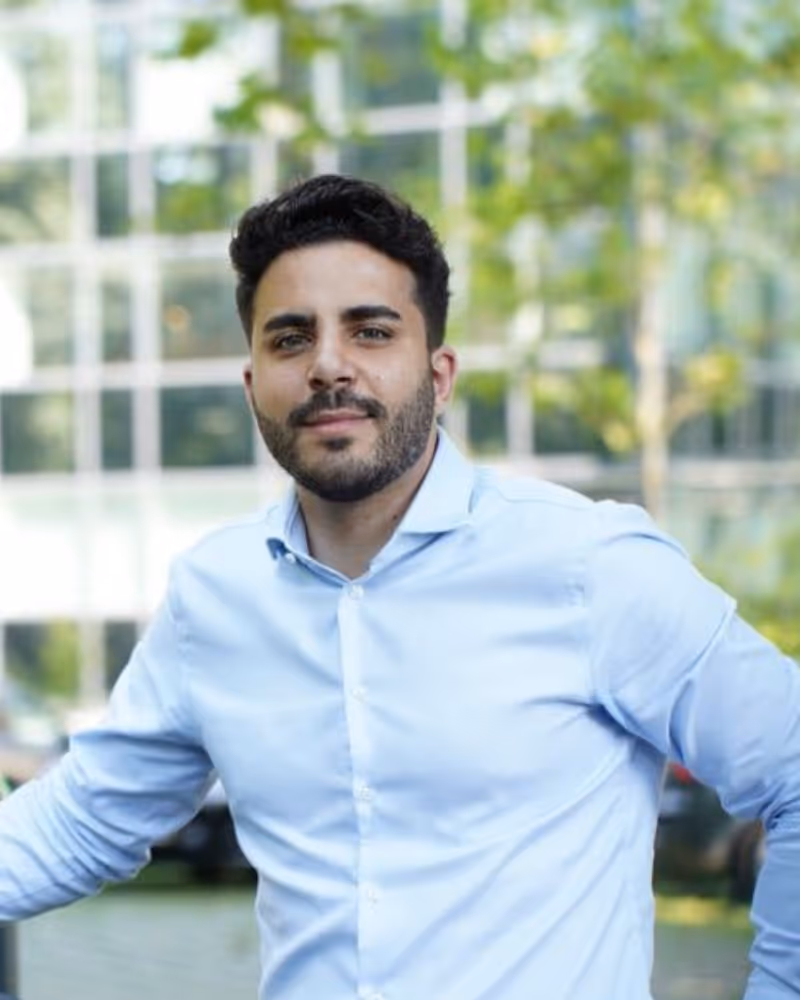 Man with dark hair and beard wearing a light blue button-up shirt, standing outdoors with blurred greenery and building in the background.