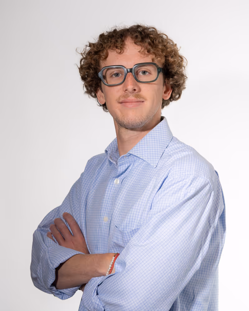 Young man with curly hair and glasses, wearing a light patterned dress shirt with arms crossed.
