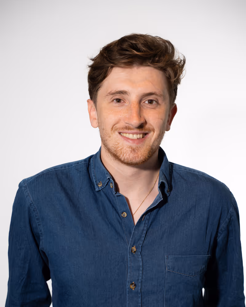 Smiling young man with short brown hair wearing a blue button-up shirt against a plain white background.
