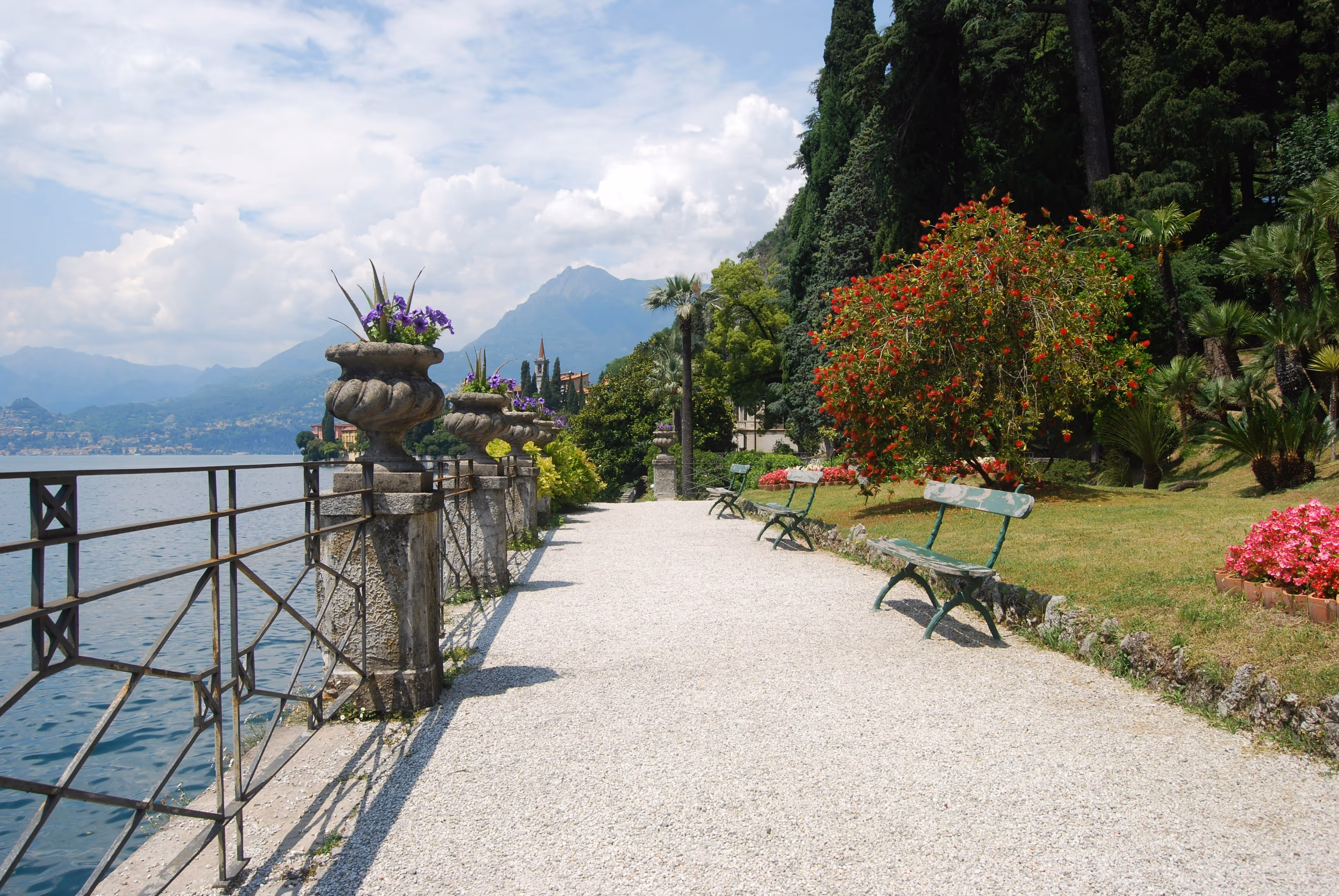 Peaceful garden walkway with benches and flowers along Lake Como, overlooking mountains and waterfront villas