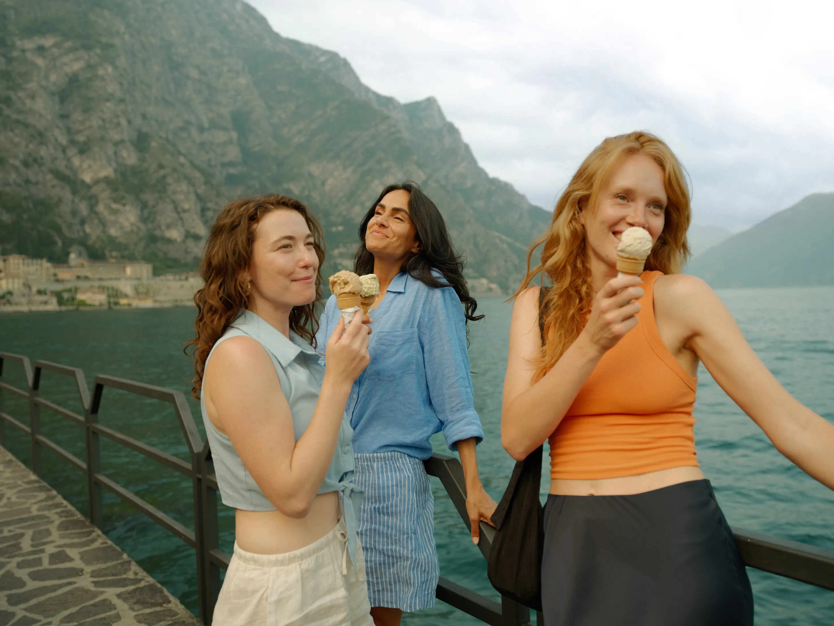 Three young women enjoying ice cream cones on a lakeside walkway with mountains in the background.