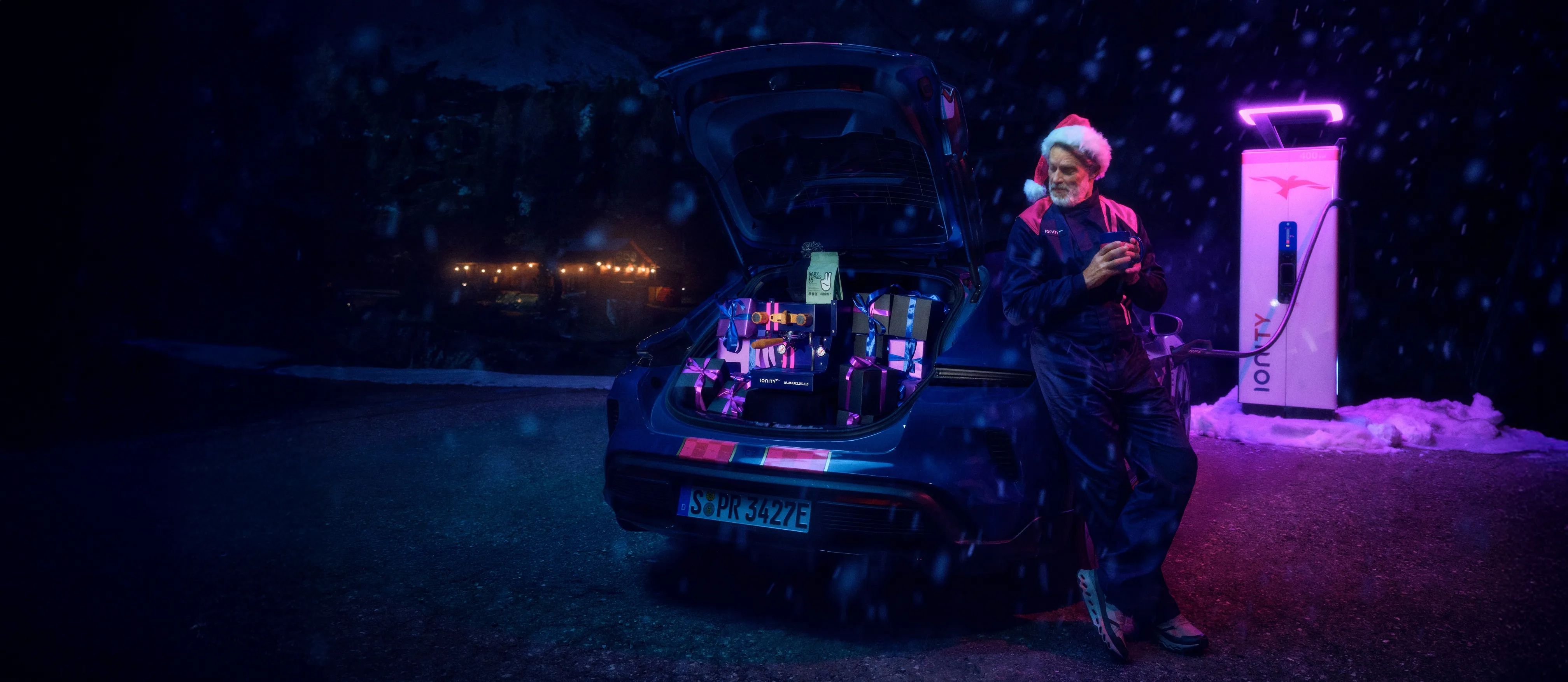 Man in a Santa hat leaning against a blue electric car with an open trunk full of wrapped gifts, next to a glowing Ionity charging station at night with light snowfall.