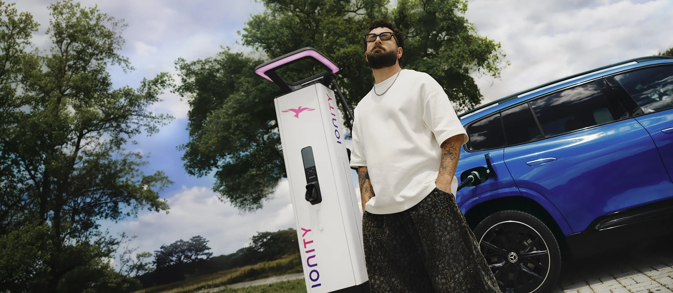 Man in sunglasses and white shirt standing by blue electric car at a white IONITY charger.