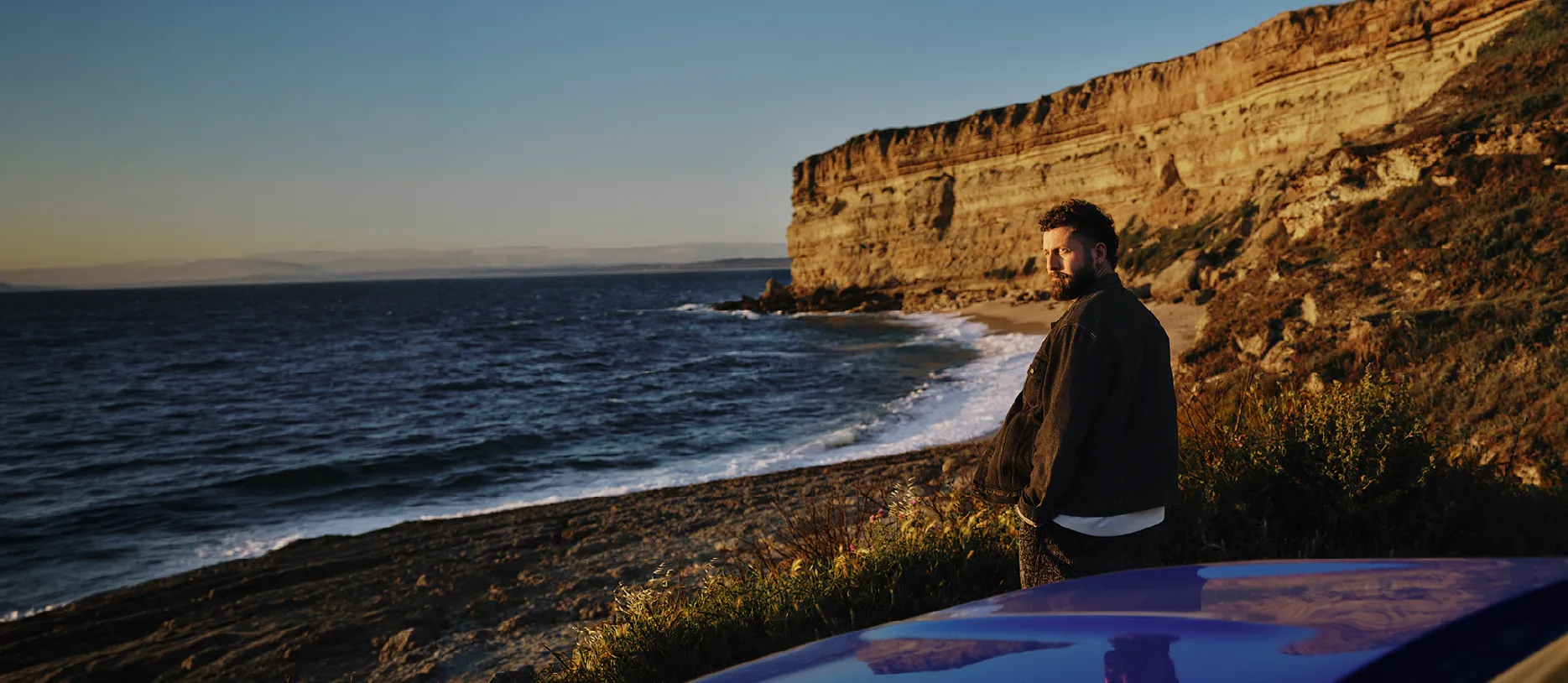 Man in dark jacket stands near a cliffside beach in front of a blue electric vehicle overlooking the ocean at sunset.