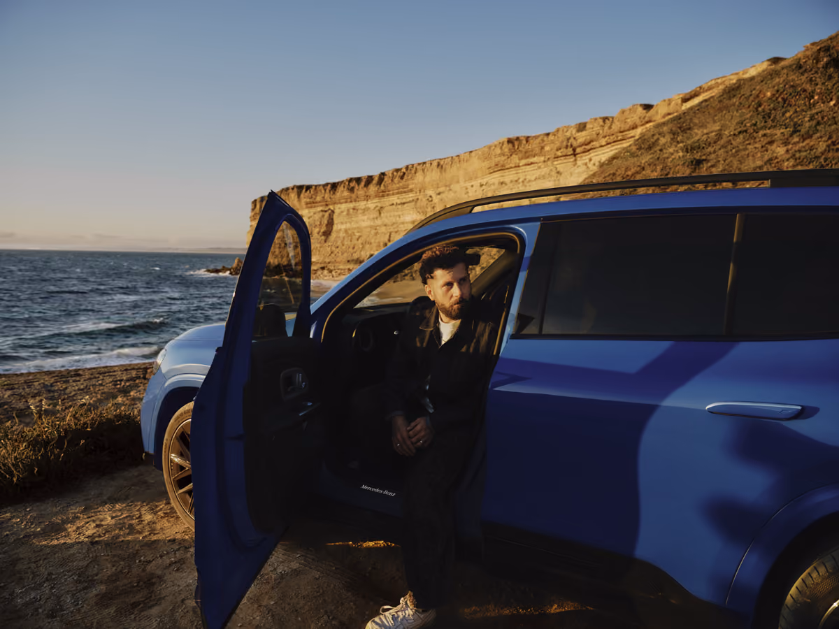 Man with beard sitting in an open blue car door near cliffs by the ocean at sunset.