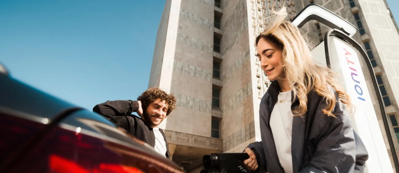 Young man and woman outdoors near parked car at an IONITY Location; woman holding the cable of an ultra-fast charger, man smiling and stretching his neck, with tall building in background under clear blue sky.