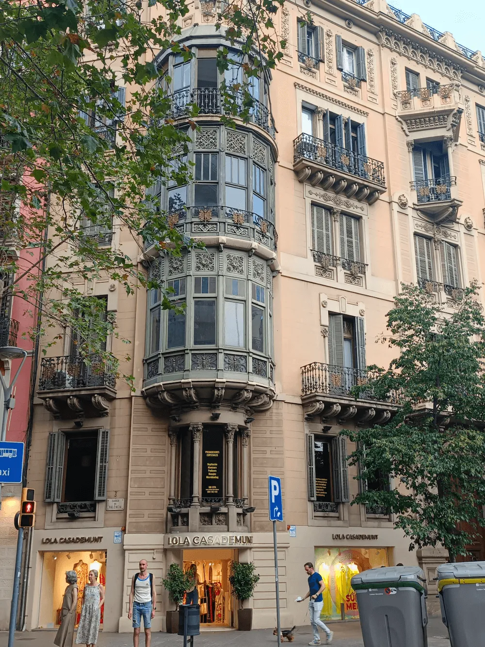 Fachada de edificio antiguo con balcones decorativos y vitrinas de tienda Lola Casademunt, personas caminando y árboles al frente.