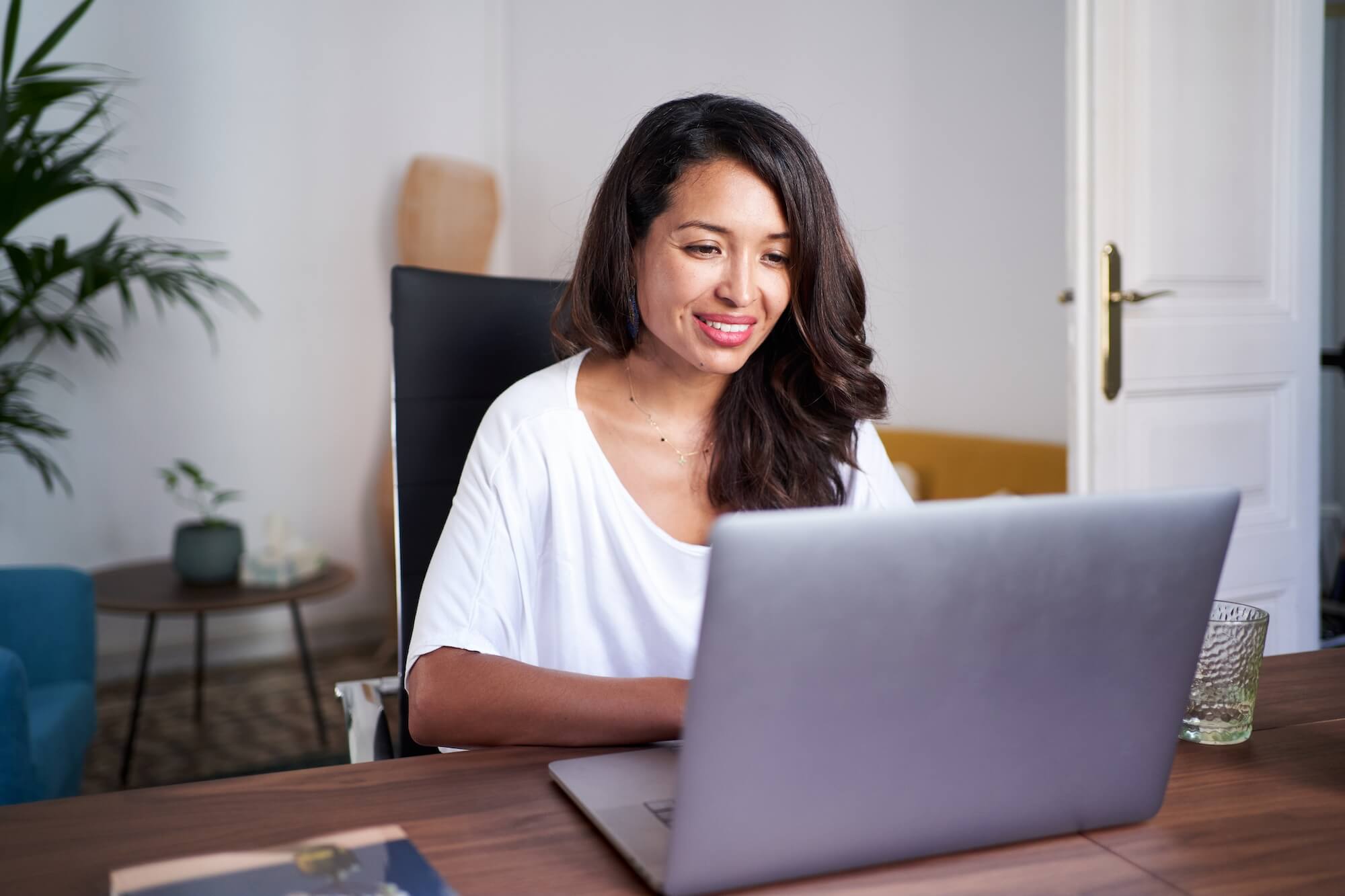 Mujer sonriente con camiseta blanca usando una laptop en un escritorio de madera en un ambiente de oficina en casa.