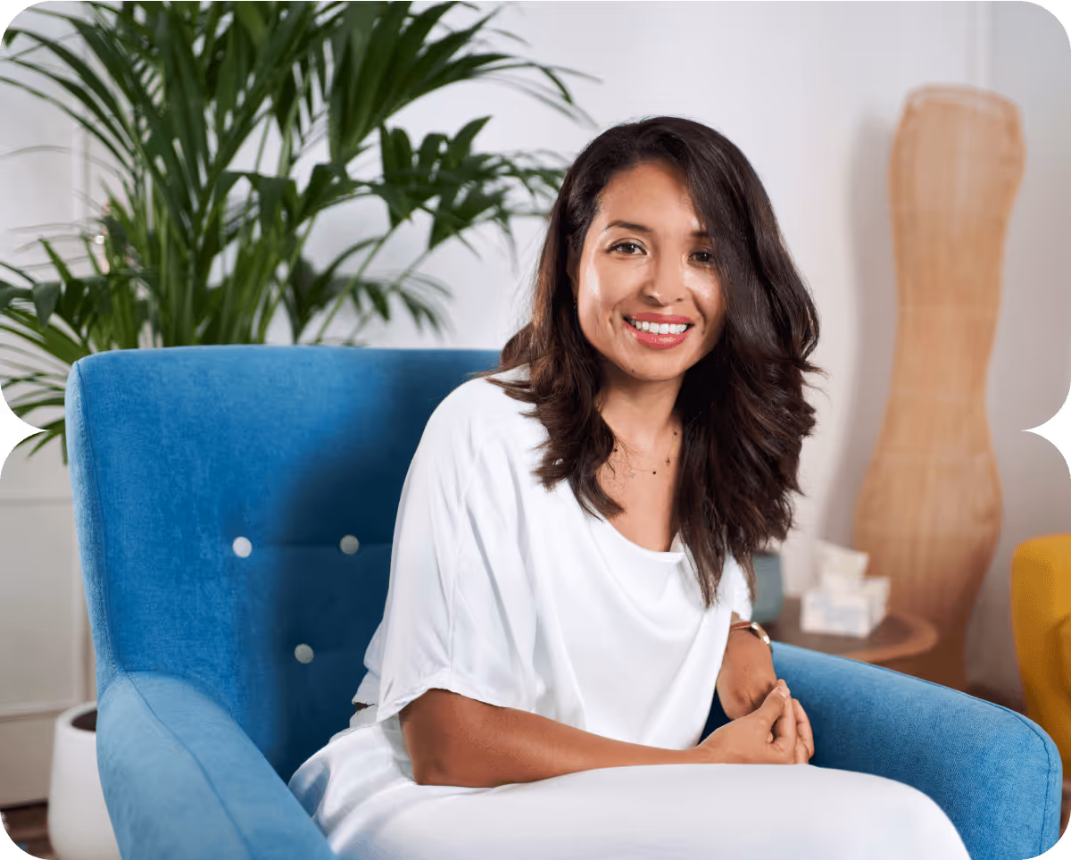 Mujer sonriente con cabello oscuro sentado en un sillón azul en una habitación con plantas y decoración moderna.