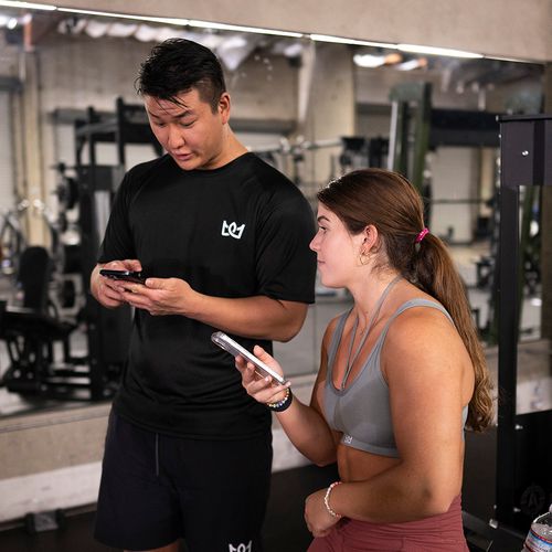 A man and a woman in workout clothes looking at their smartphones in a gym with exercise equipment and a mirror behind them.