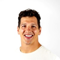 Smiling man with dark curly hair wearing a white shirt against a white background.