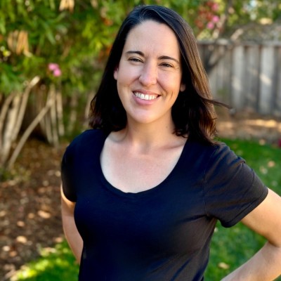 Smiling woman with dark hair wearing a black shirt standing outdoors with greenery and a wooden fence in the background.