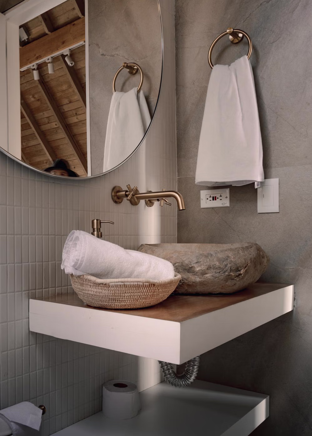 Lavamanos de piedra con grifería dorada, toalla blanca y espejo redondo en baño moderno.