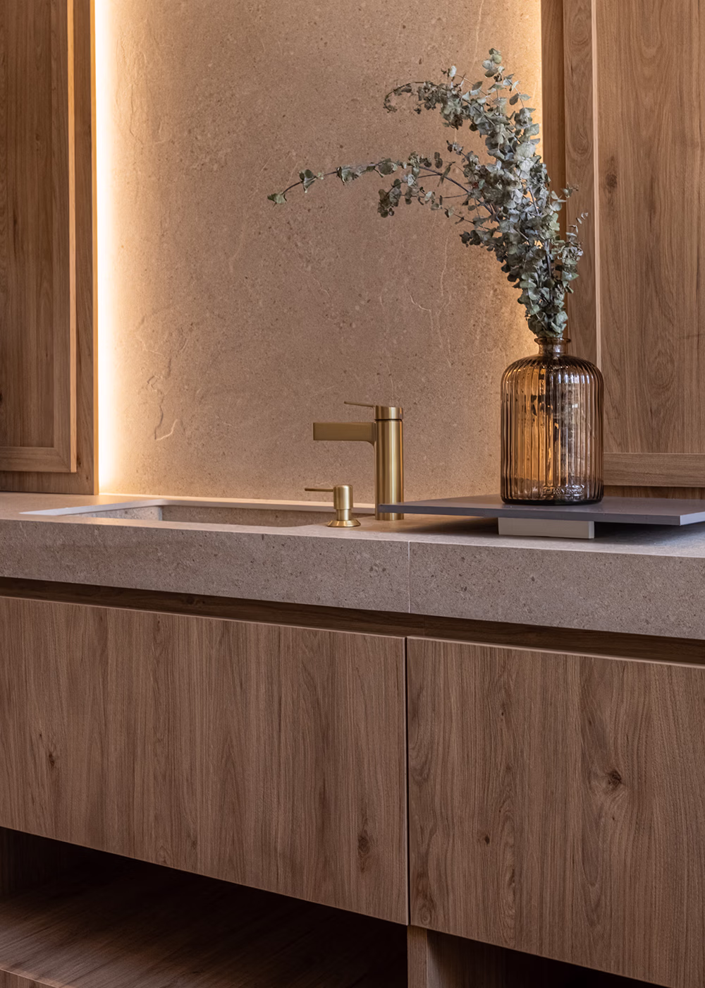 Lavabo moderno con mueble de madera, grifería dorada y jarrón con ramas secas sobre la encimera.