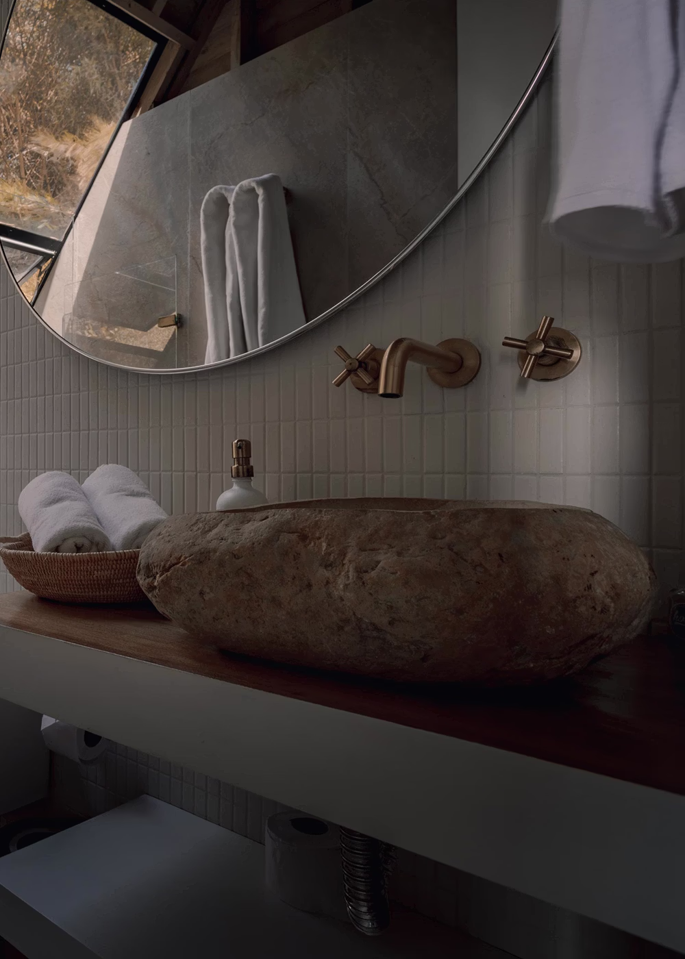 Lavabo de piedra y grifería dorada con espejo redondo y toallas blancas en baño moderno.