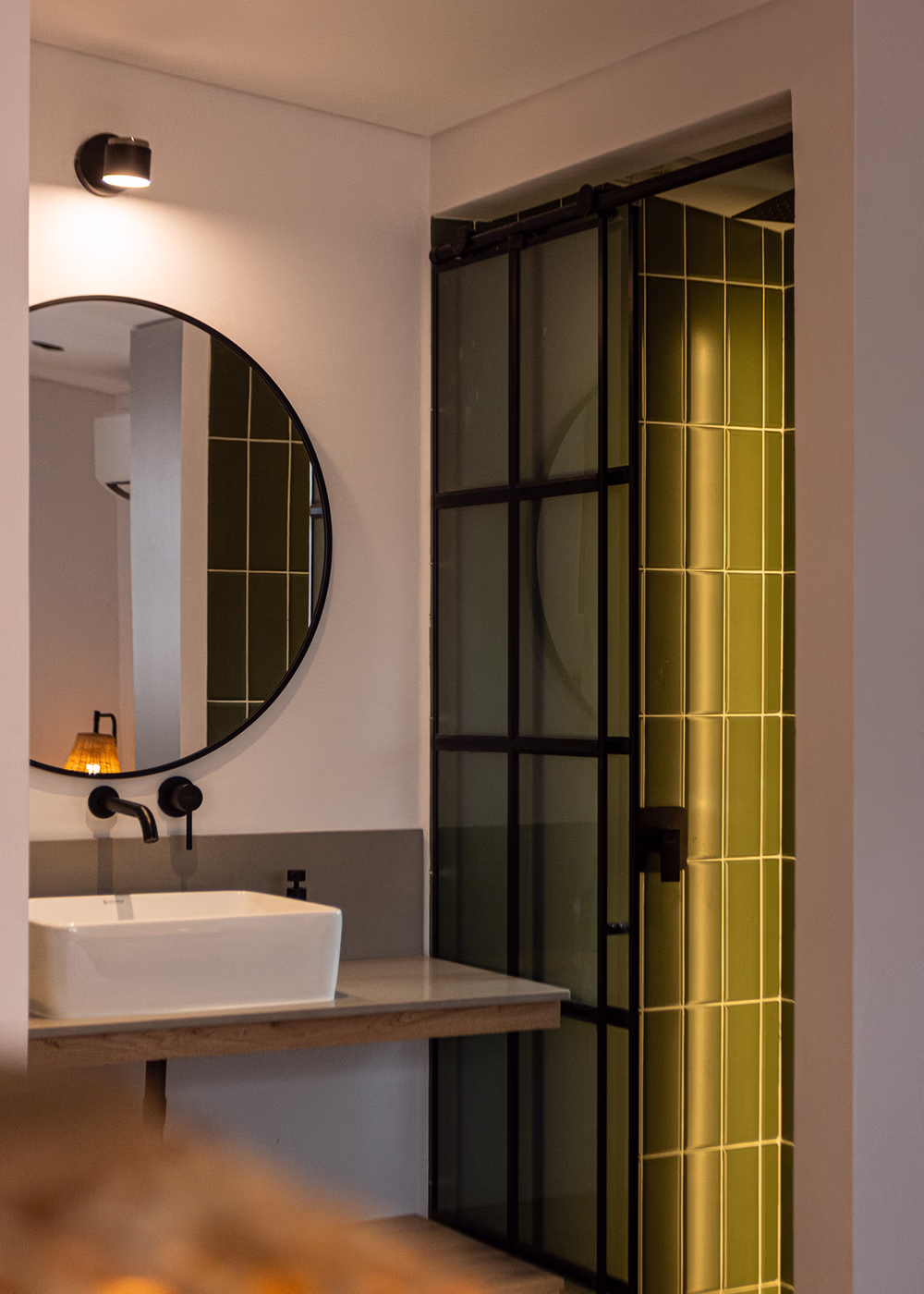 Baño moderno con lavabo blanco, espejo redondo y ducha con puerta de vidrio y azulejos verdes.