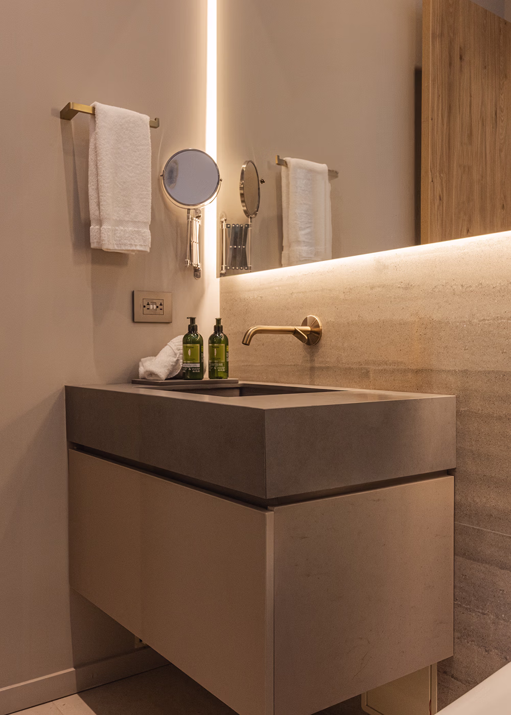 Lavamanos moderno con toalla blanca, espejo de aumento y dispensadores verdes en baño beige.