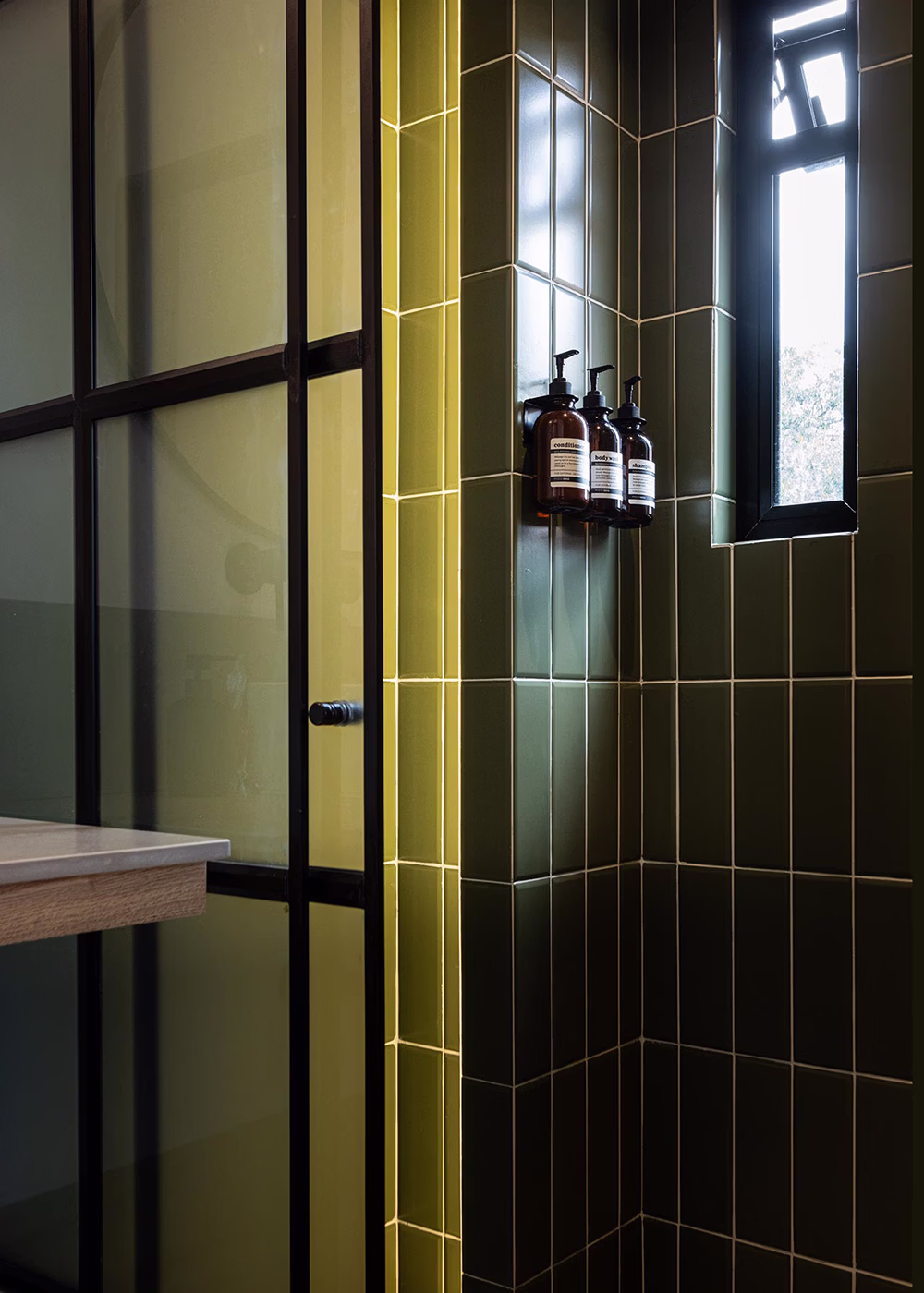 Esquina de ducha con azulejos verdes, ventana pequeña y dispensadores de champú, acondicionador y gel.