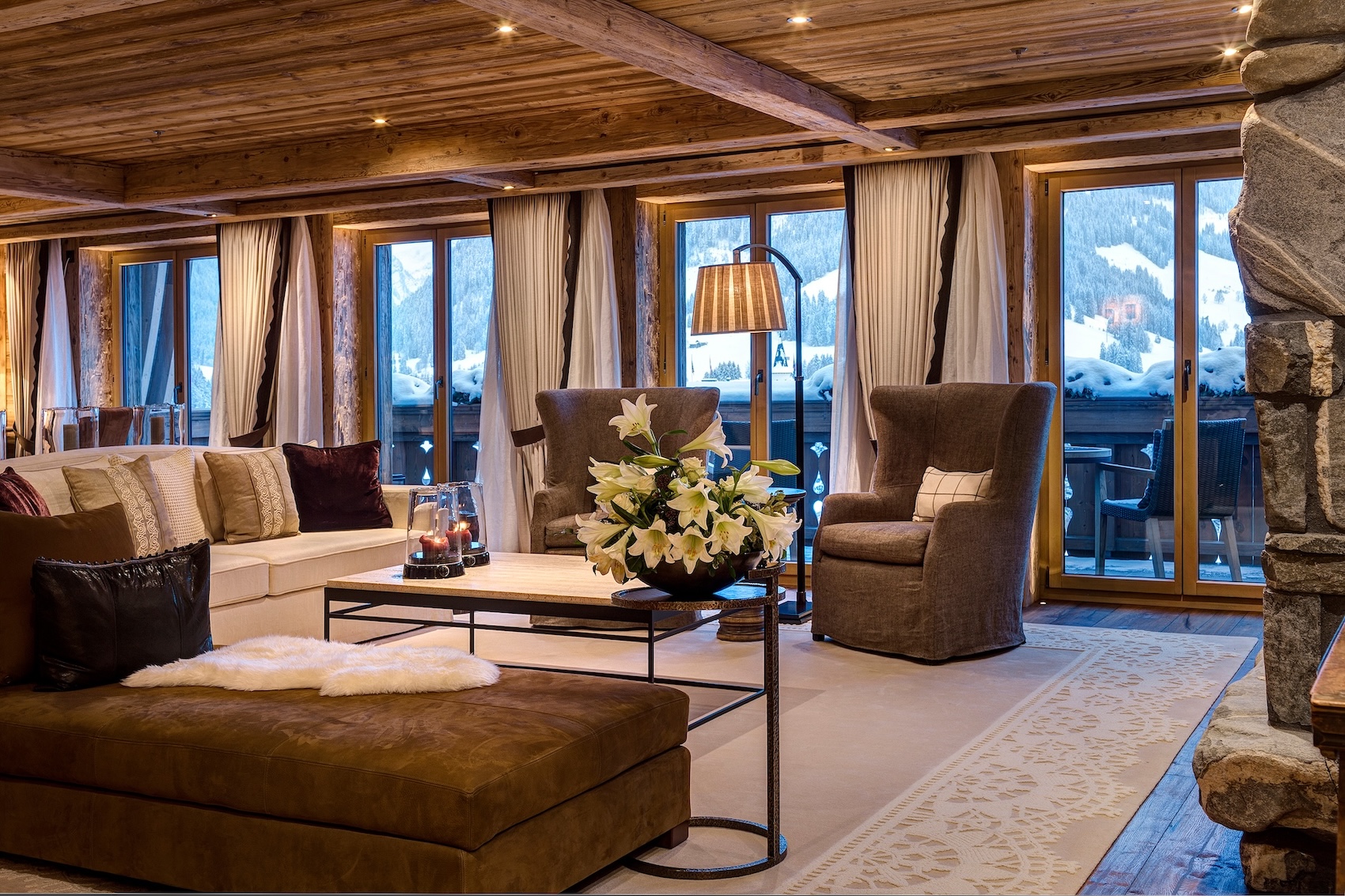 The Panoramic Residence living room at The Alpina Gstaad, with timber-beamed ceilings, a stone fireplace and floor-to-ceiling windows overlooking the snow-covered village of Gstaad.