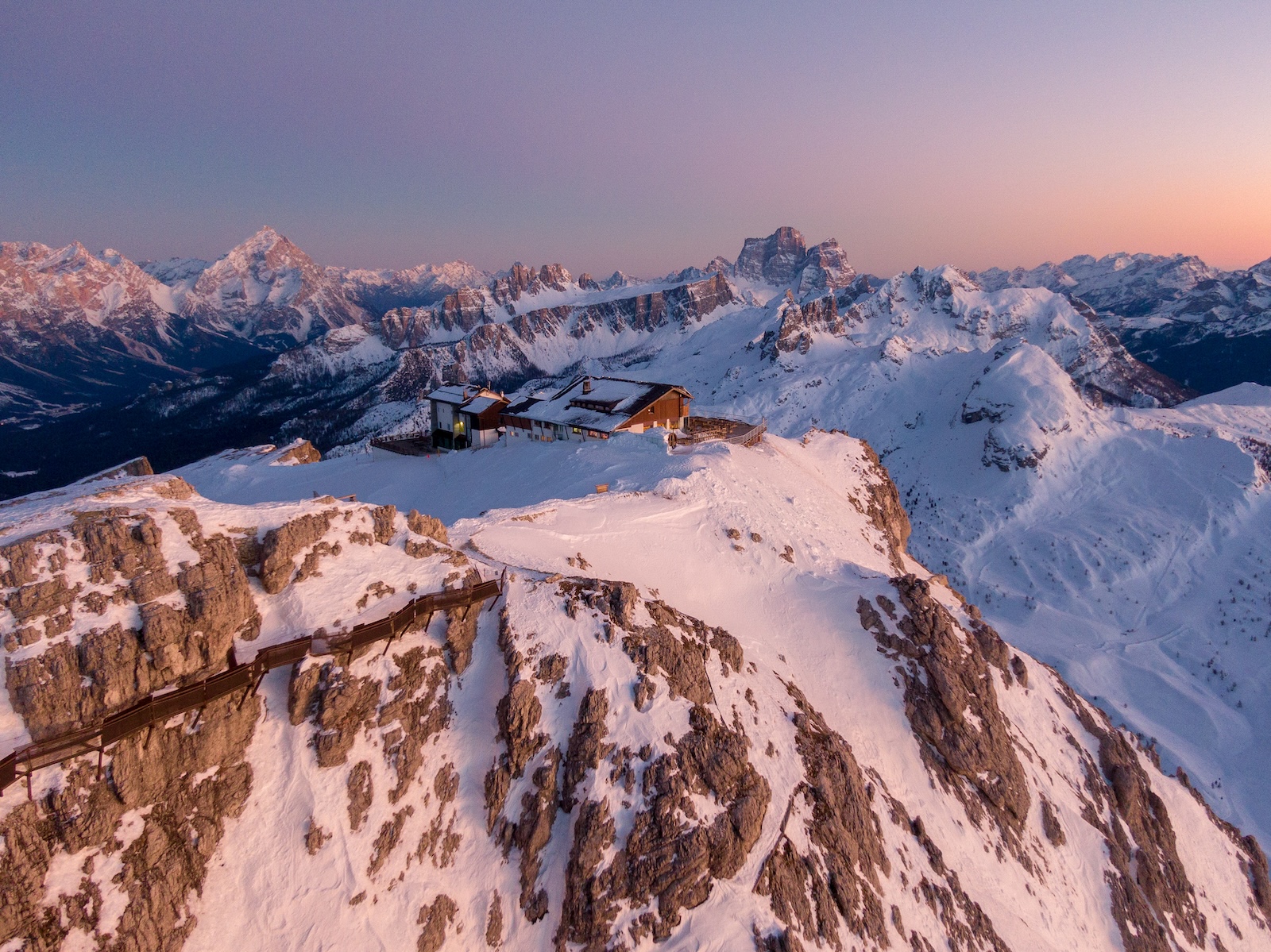 A rifugio perched on a rocky Dolomites summit at dusk, its windows lit against a pink alpenglow sky, with layered snow-covered peaks receding into the distance