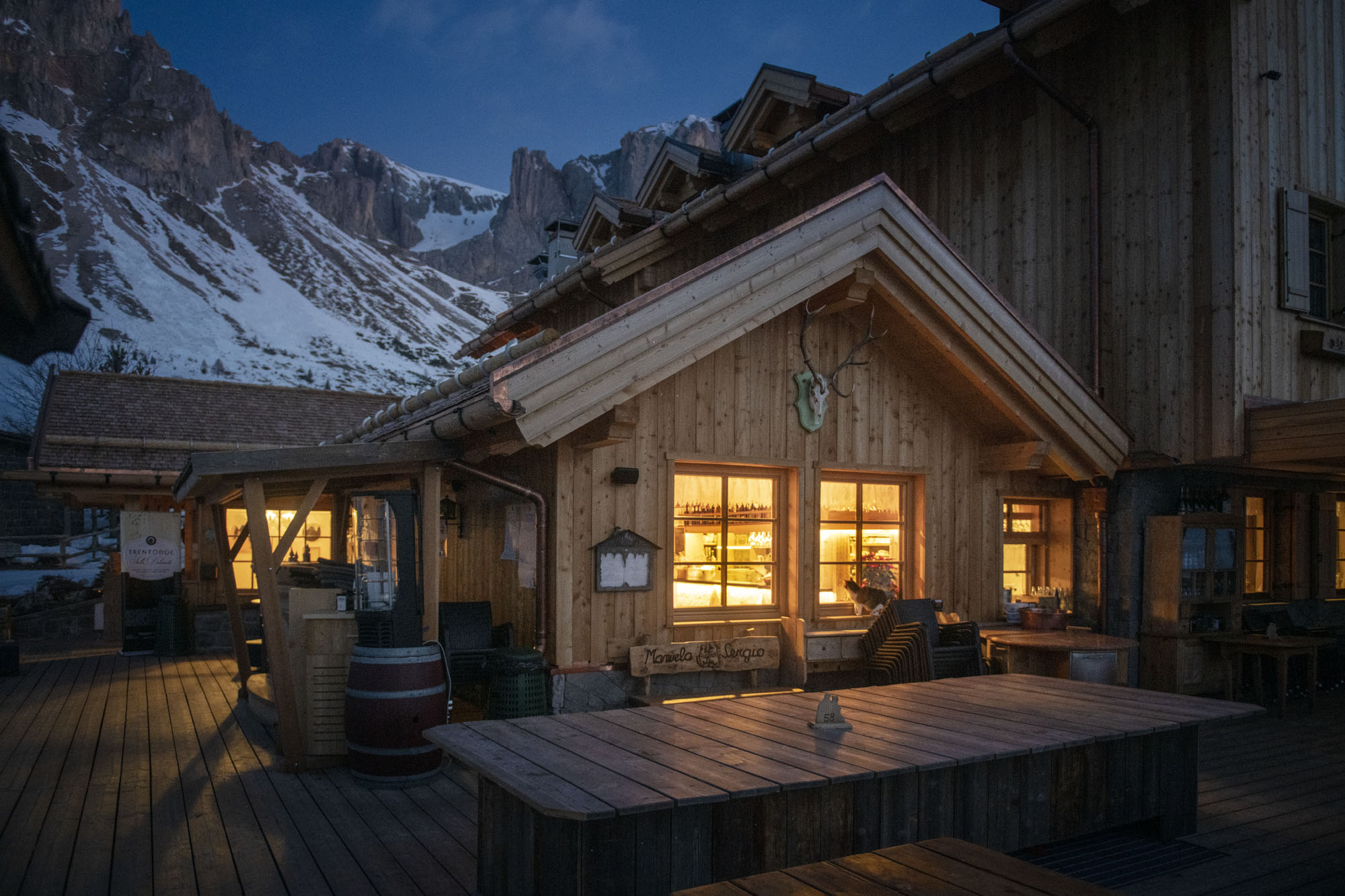 A timber mountain chalet with warmly lit windows on a wooden terrace at dusk, jagged snow-dusted peaks rising behind against a darkening sky.
