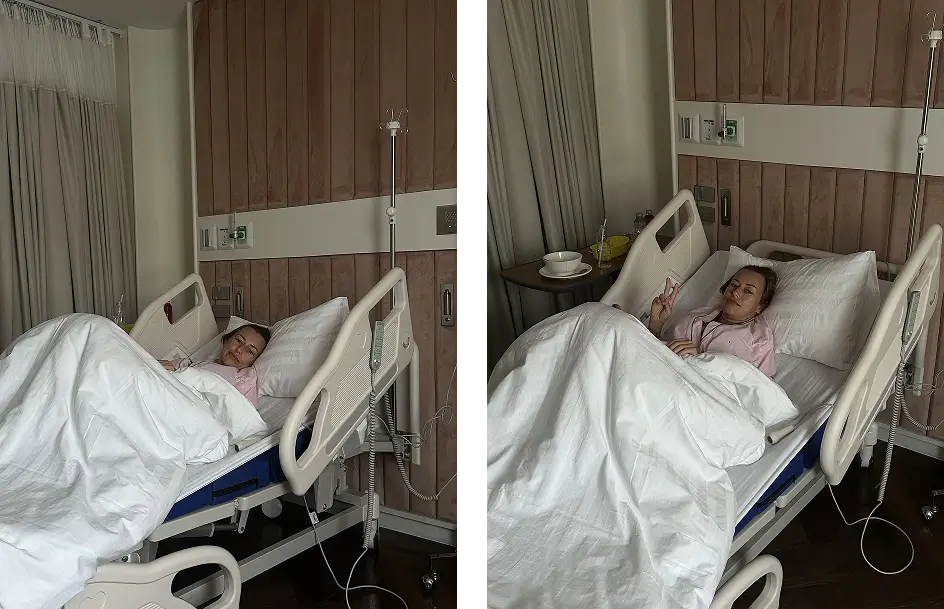 Julia resting in her hospital bed after breast lift with implants surgery in Bangkok, smiling and making a peace sign