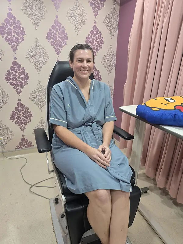 Racheal sitting in a pre-op chair at World Medical Hospital, wearing a blue hospital gown, smiling calmly before her surgery.