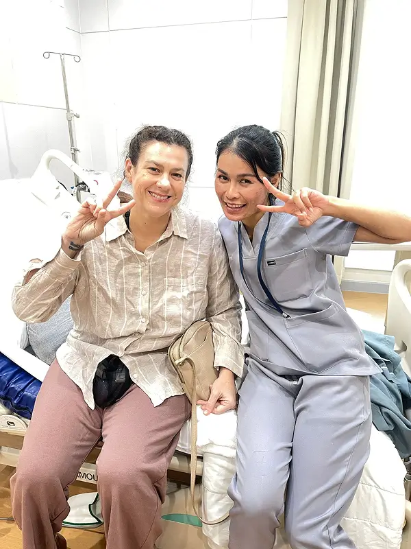 Racheal sitting on her hospital bed post-surgery, smiling with a Beauty Butler nurse in blue scrubs beside her.