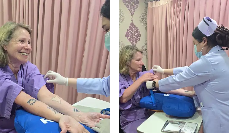Lori smiling during pre-surgery preparation as a nurse marks her arm and prepares the IV at World Medical Hospital, Bangkok.