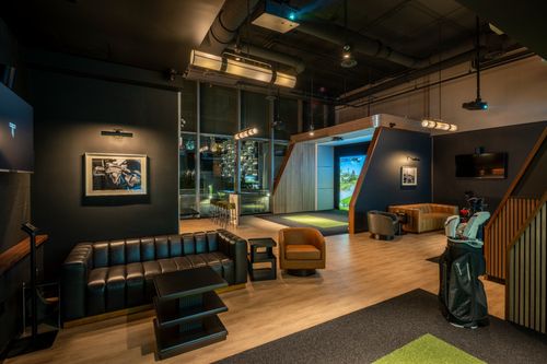 Lounge seating area opening to a golf simulator bay; golf bag and modern lighting visible.