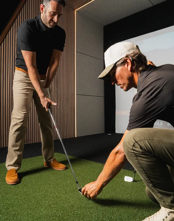 Instructor kneels to align the clubface while a student watches during an indoor golf lesson.