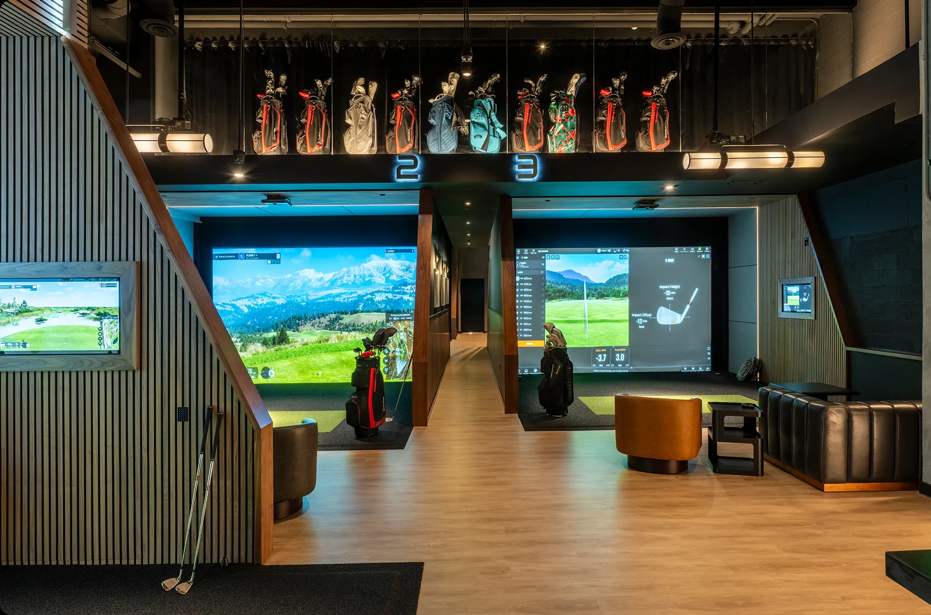 Modern indoor golf studio with two simulator bays, lounge seating, and golf bags displayed above the bays.