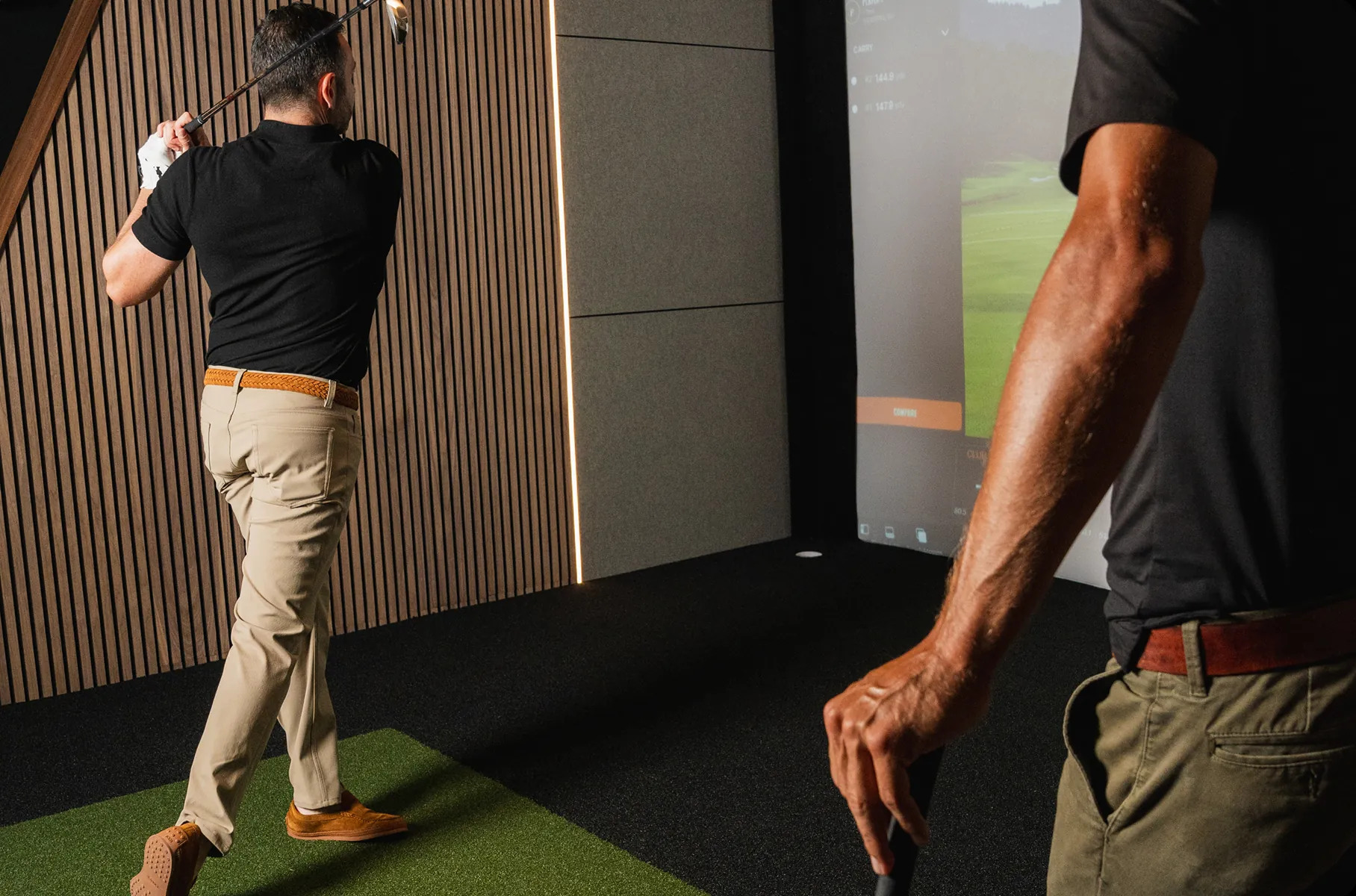 Indoor golf lesson—one player finishes a swing toward the simulator screen while the coach watches in the foreground.