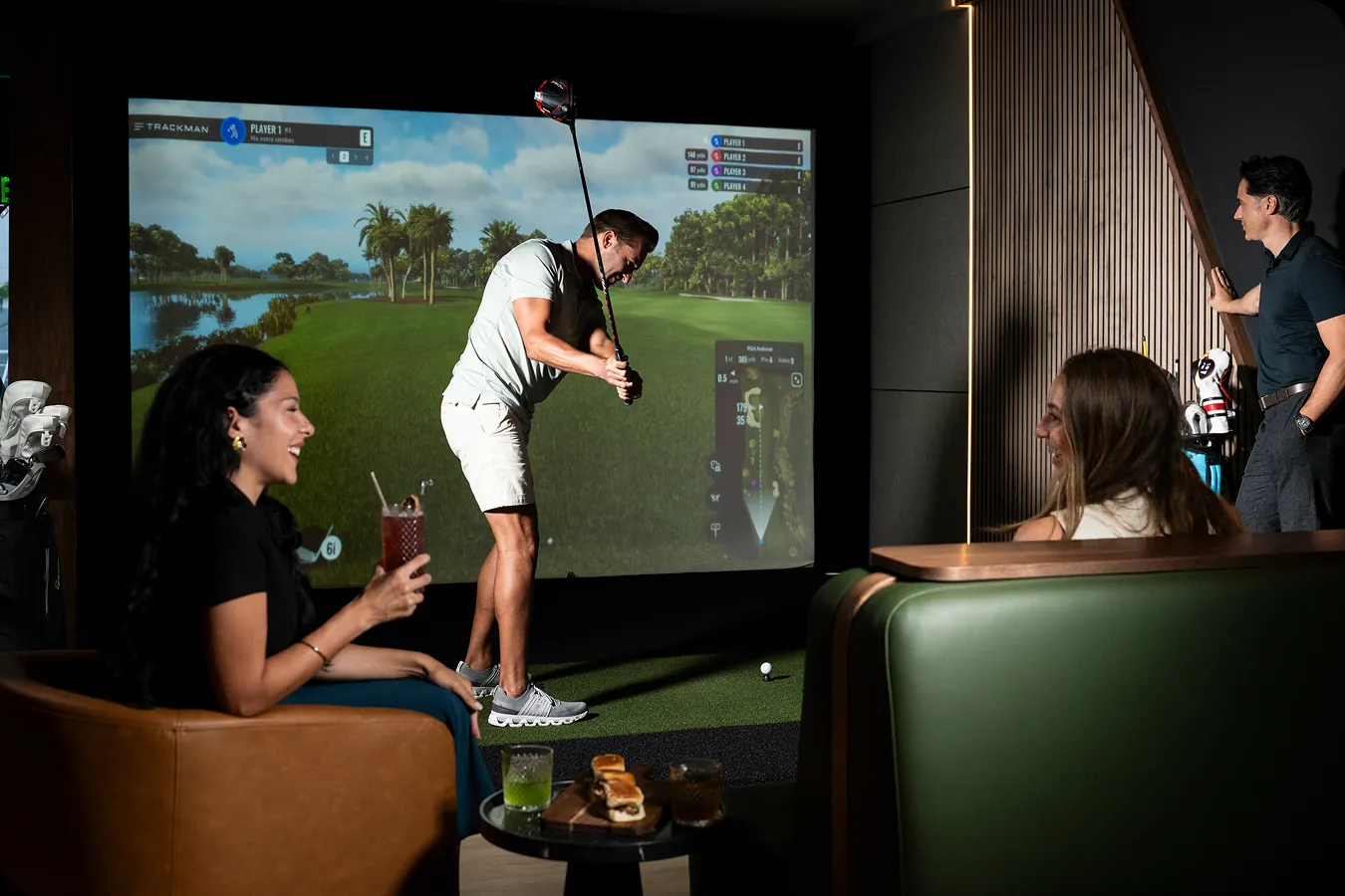 Friends relax with drinks while a golfer swings in an indoor simulator bay.
