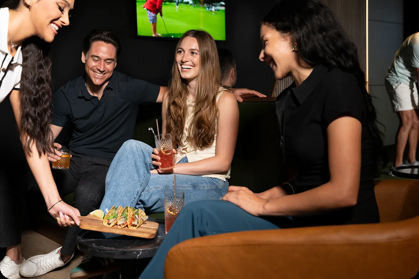 Group laughing on lounge seating as a server sets down a board of tacos and cocktails.