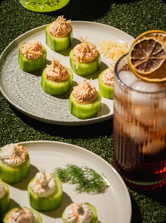 Plate of cucumber rounds topped with herbed filling beside a green basil cocktail on turf.