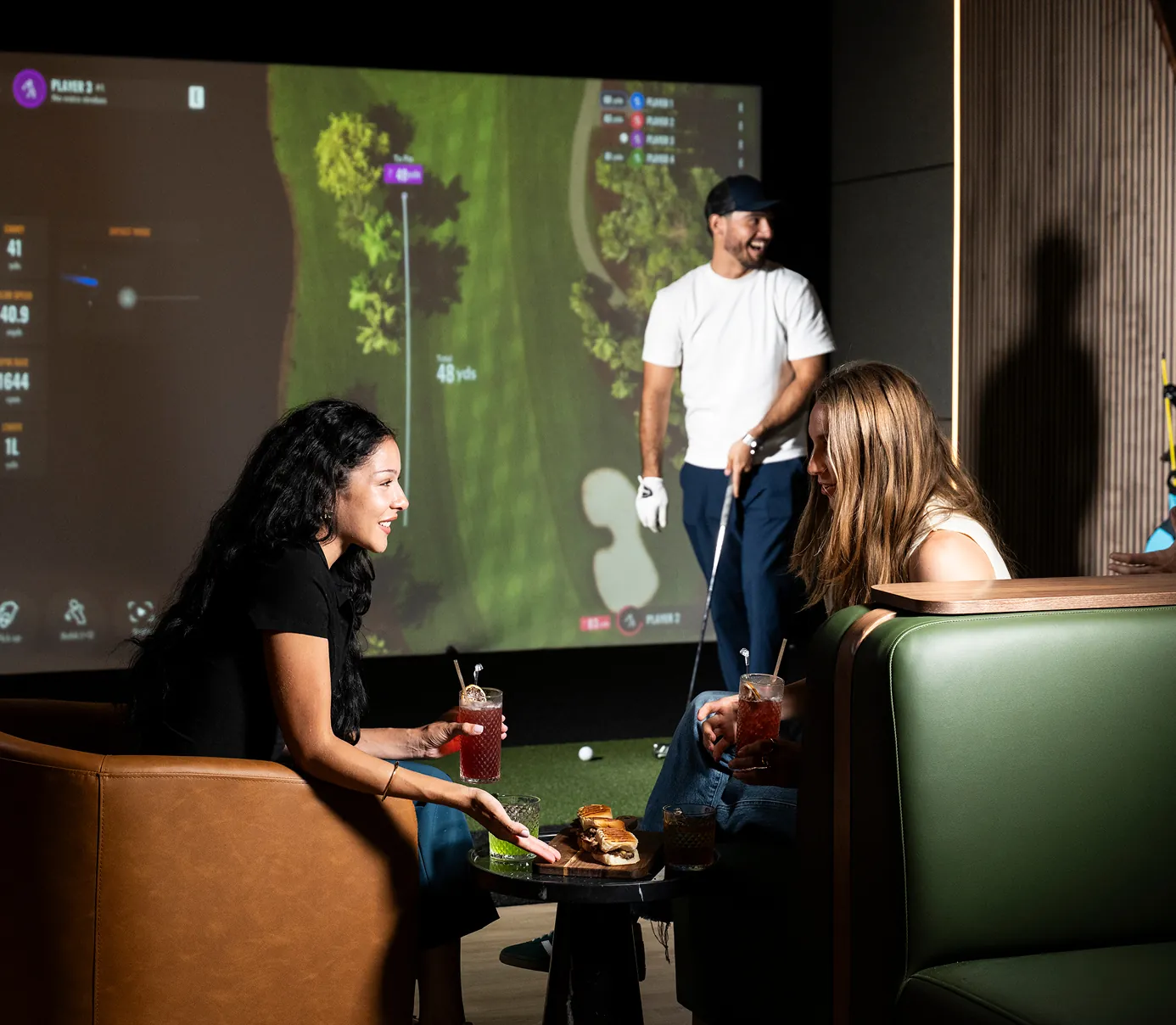 People socializing inside a Tips modern indoor , golf simulator lounge, two women chatting on sofas with drinks while a man stands in the simulator bay.